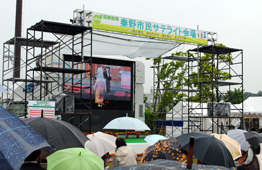 雨天の屋外で、鉄骨で組まれたやぐらの中央に大型ビジョンが設置され、上部には「秦野市民サテライト会場」と書かれた横断幕があり、手前には傘をさした大勢の観客が集まっている写真