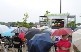 雨が降る屋外イベント会場で、様々な色や柄の傘をさした観客が椅子に座って、鉄骨で組まれたステージに設置された大型ビジョンでの映像を視聴している写真