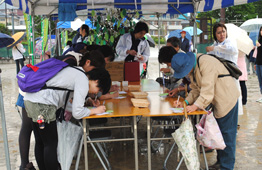 雨の屋外イベント会場で、青と白のストライプのテントの下にあるテーブルを大勢の参加者が囲み、それぞれが手書きで緑色の葉の形をした札のようなものに記入している様子の写真