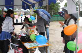 雨が降る屋外で、青と白のストライプのテントの下にある受付のようなブースで、女性が来場者とやり取りをしている写真