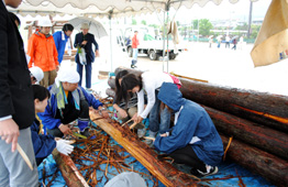 雨の屋外イベントのテントの下で、参加者たちが丸太の周りでかがみ込み、丸太切り体験をしている写真