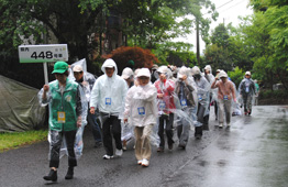 プラカードを掲げたスタッフが、透明なカッパを着用した参加者たちを植樹会場へ案内している写真
