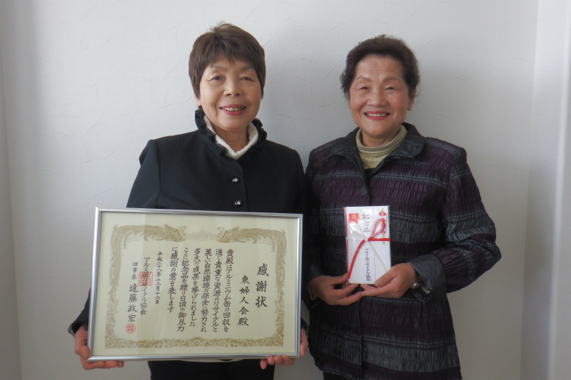 白い壁を背景に、感謝状を掲げた女性と、熨斗袋を手に持った女性が並んで笑顔を見せている写真