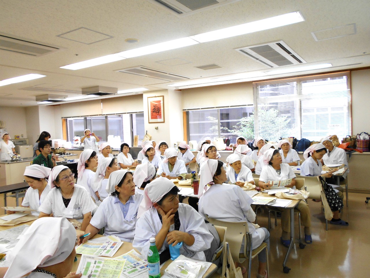 白い制服と三角巾をつけた複数の女性たちが、資料を広げた机の席に座って、熱心に講師の話を聞いている様子を撮影した写真