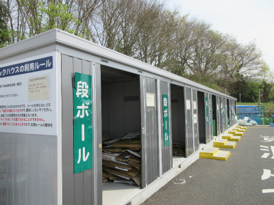 広々とした駐車場の一角に、リサイクル品目ごとに仕切られた横長の金属製ボックスの集積所があり、手前の二つの扉には「段ボール」と大きく緑色で表示された案内板が貼られ、その奥にも同様のボックスが並んでいる環境資源センターのストックハウスの写真