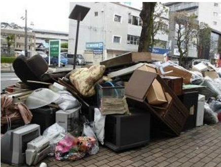 街路樹のそばの歩道上に、木製家具、水槽、テレビ、パソコン、ストーブ、ごみ袋などが雑多に山積みになっている、ごみ収集場所に出された片付けごみの写真