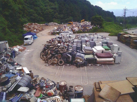 山間部の広大な屋外の集積所で、大量の古タイヤ、冷蔵庫や洗濯機などの大型家電、マットレス、金属くず、廃材、ドラム缶といった様々な種類の廃棄物が種類別に積み重ねられており、奥には重機と白いトラックが見える、粗大ごみが分別され整理された仮置場の写真