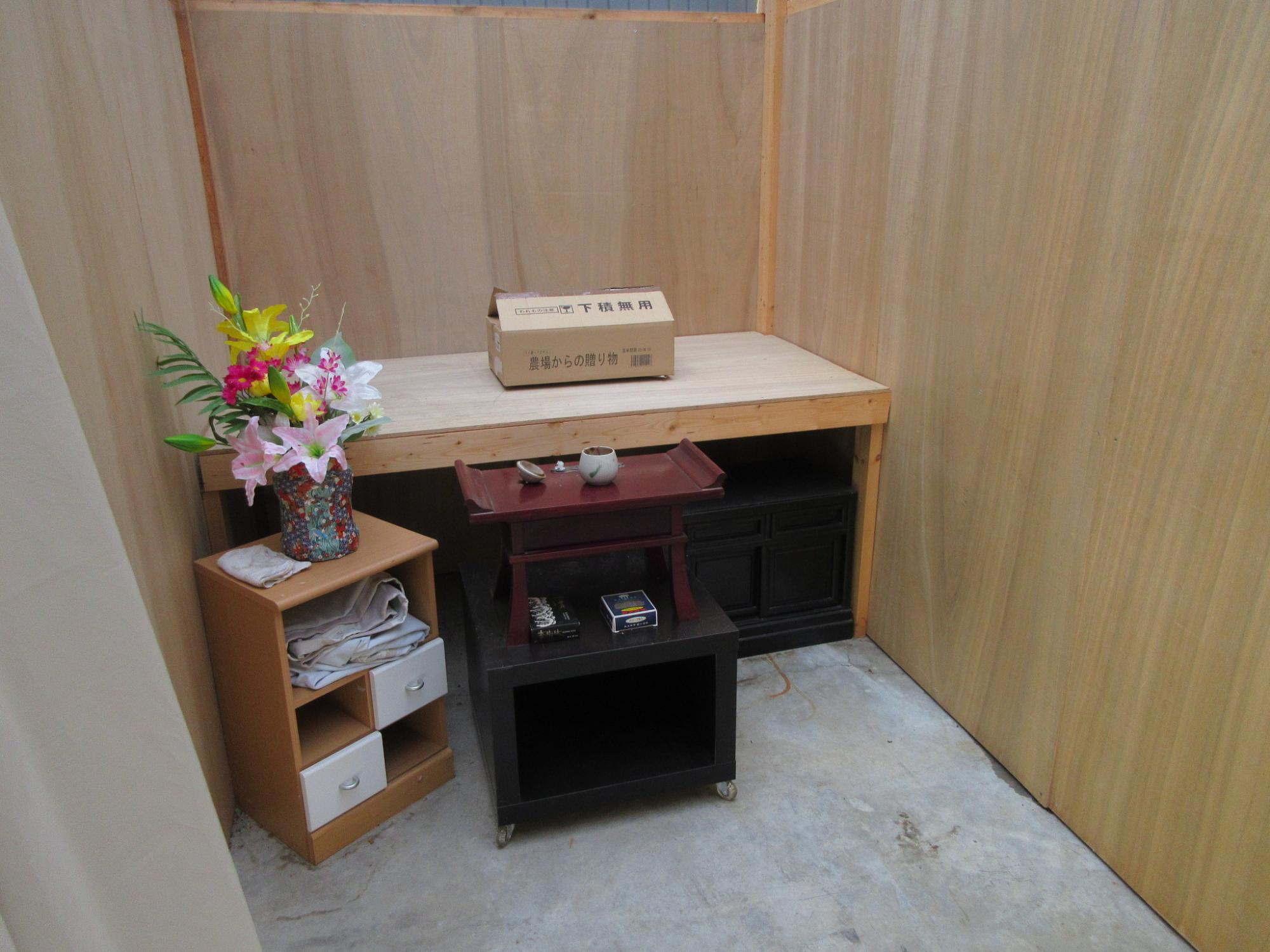 簡易なベニヤ板のような壁で囲まれた室内の一角で、黒い台と、供花、線香立て、台の上に段ボール箱が置かれている一時安置所の写真