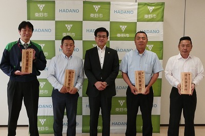 秦野市のバックパネルの前で、市長を中央に木製の看板を持った代表者の男性4名が横一列に並んでいる記念写真