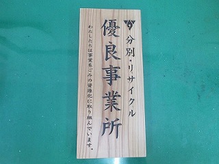 分別のリサイクルに取り組んでいることを示す、「優良事業所」と縦書きされた文字が中央に書かれ、左に「わたしたちは事業系ごみの資源化に取り組んでいます。」という小さな文字が添えられている木製の立て札のような看板が緑色の台の上に置かれている写真