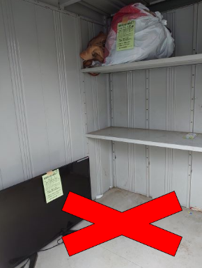 ストックハウス内の床に大型の薄型テレビが置かれ、上段の棚には衣類のようなものが入った袋が置かれており、赤い大きなバツ印が重ねて表示されている写真