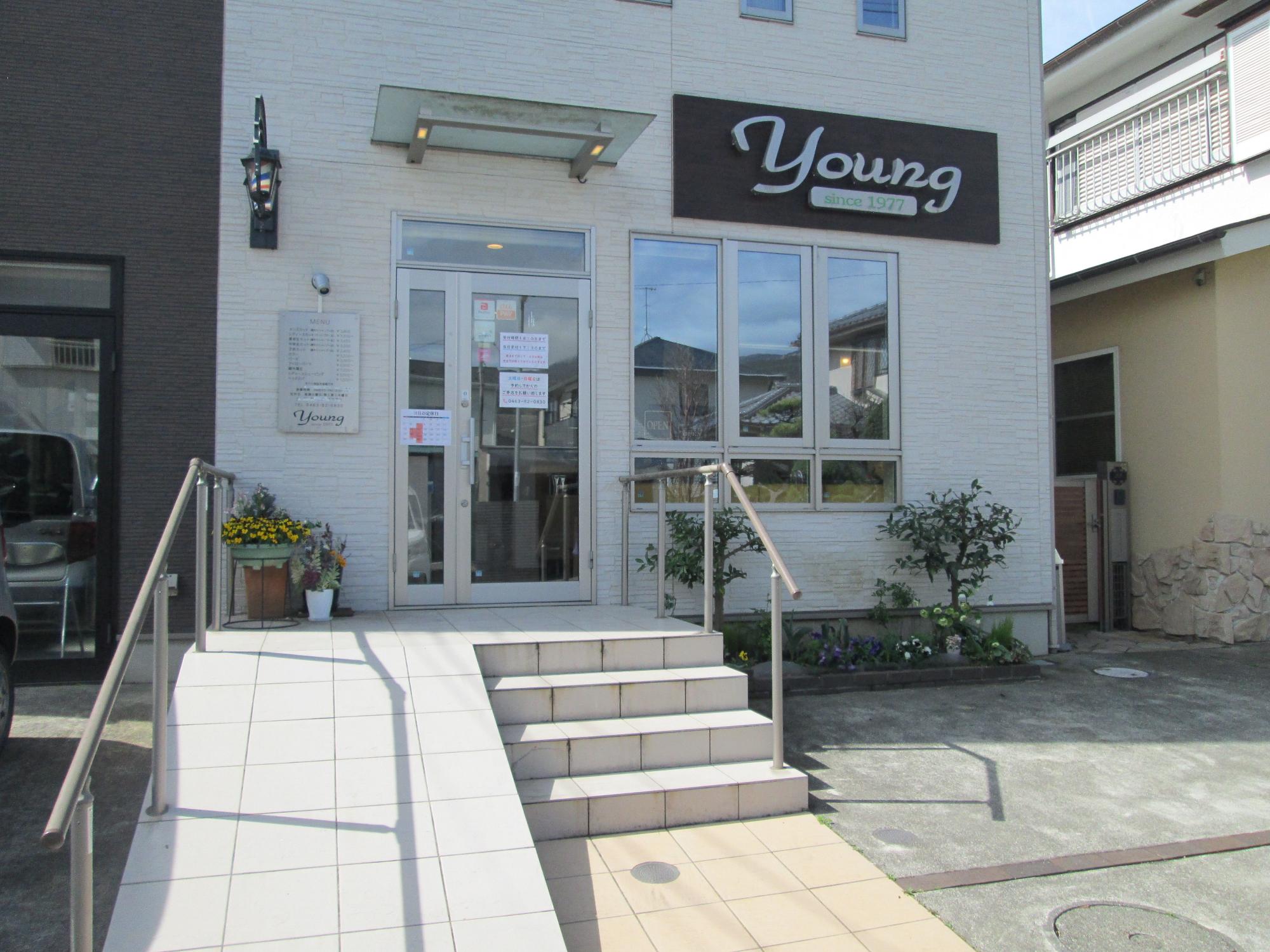 タイル張りのスロープと階段が設置された、白い壁に「Young」と書かれた大きな看板取り付けられている店舗の入り口前の写真