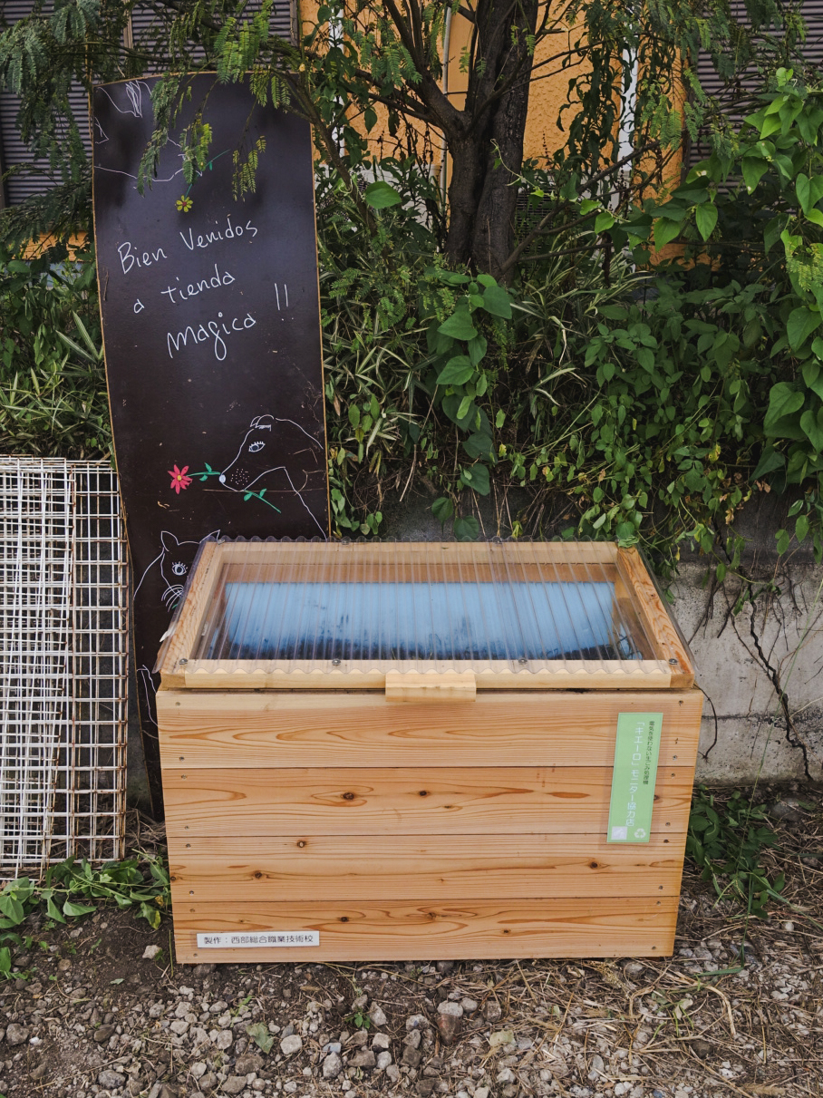 屋外の植え込み前に置かれた、「Bien Venidos a tienda Magica !!」と書かれた細長い黒板の前に設置されたキエーロの写真