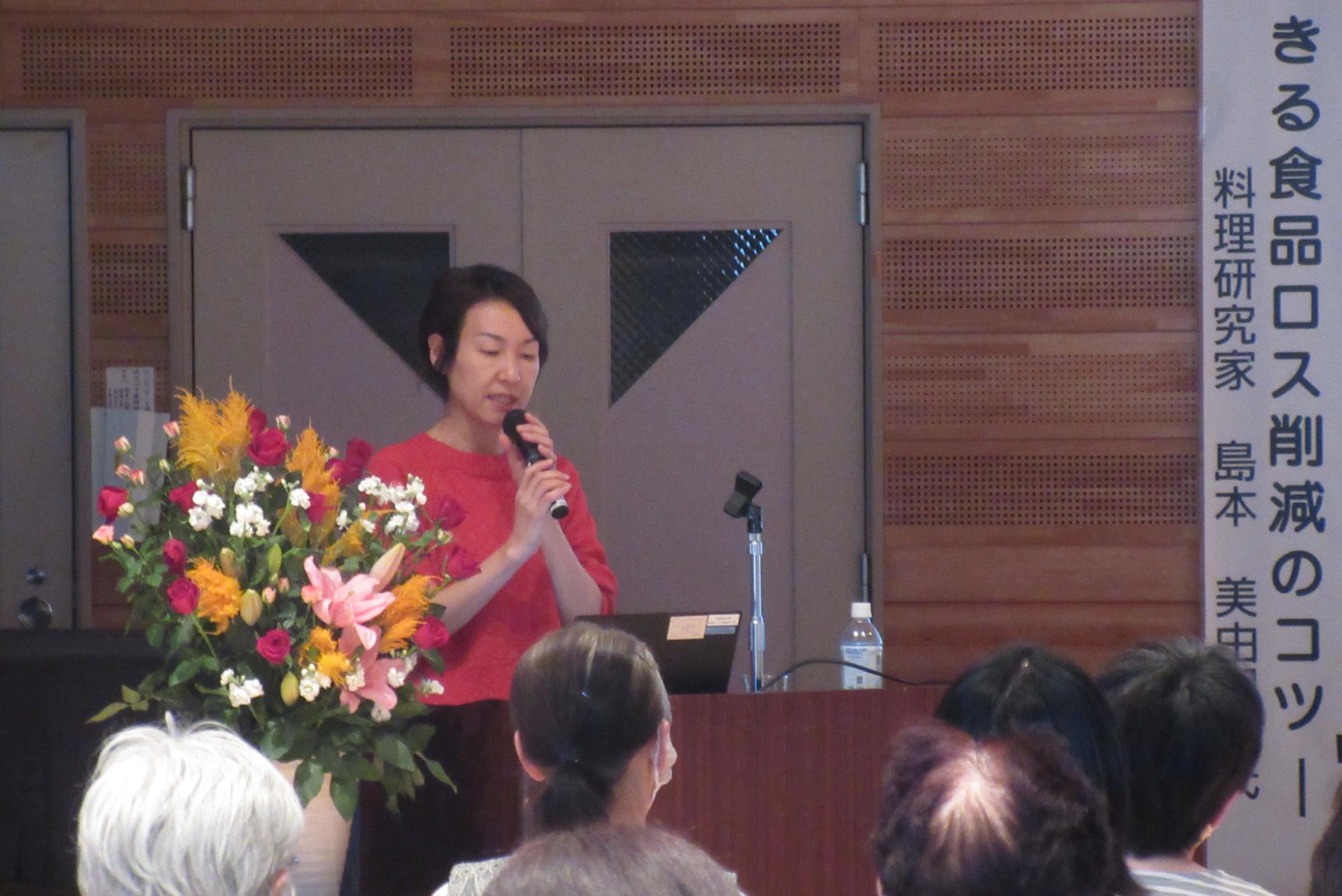 会場のステージ上で、料理研究家の島本美由紀氏が、豪華な花が飾られた演台でマイクを持ち、聴衆に向けて「食品ロス削減のコツ」に関する講演をしている様子の写真