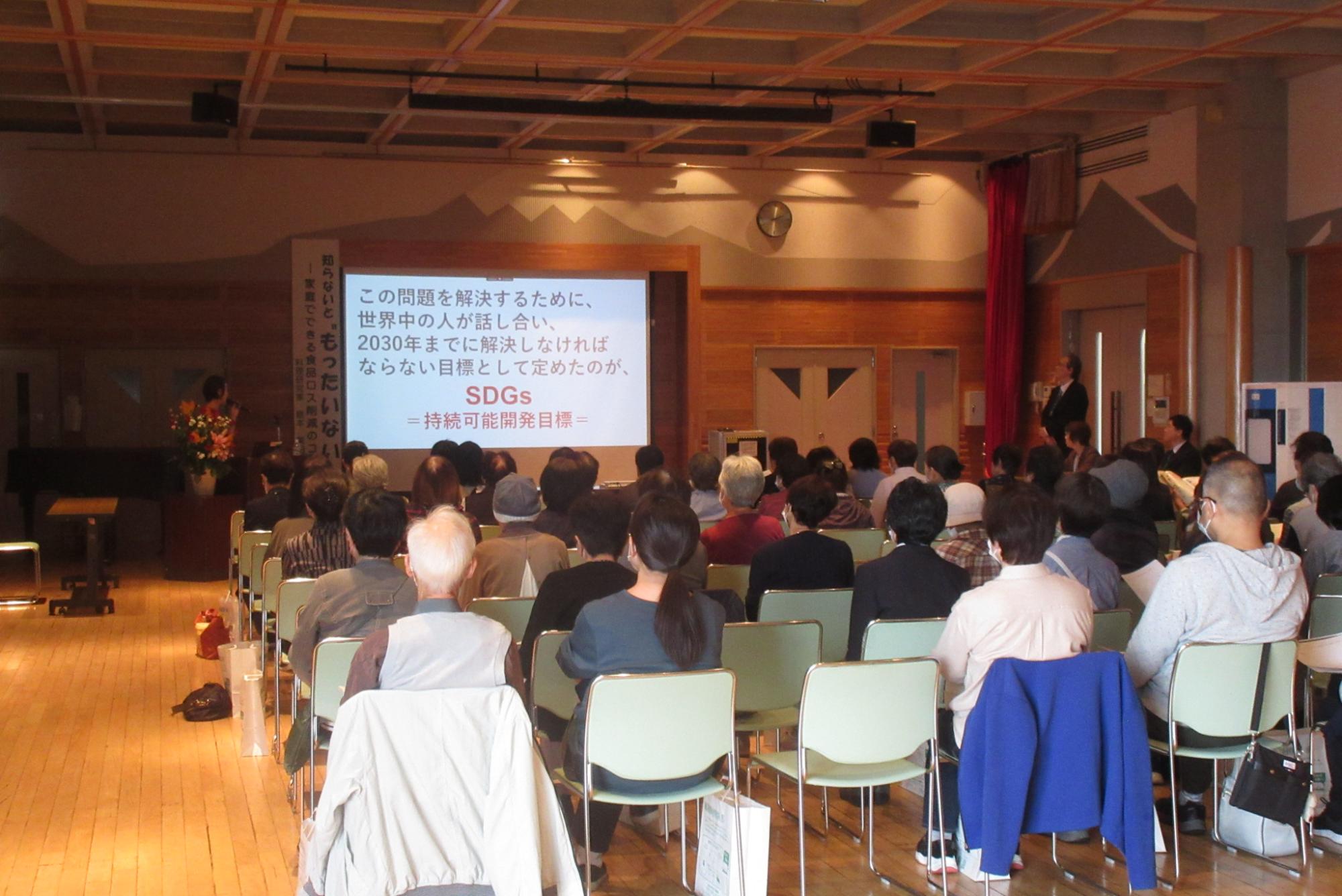 広い会場で、大勢の参加者が椅子に座って前方の資料が映し出されたスクリーンを見ている、講演会風景の写真