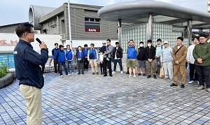 青いジャンパーを着たグループとカジュアルな服装のグループの約二十人の前で、市長がマイクを持って挨拶をしている、オリエンテーションの様子の写真