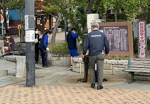 屋外で、四人の人々が清掃活動を行っており、一人がロゴ入りジャンパーを着て手石造りの構造物に近づき、他の三人は軍手やゴミ袋を持って地面のゴミを拾っている地域清掃ボランティアの様子の写真