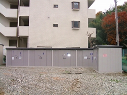 大きな集合住宅の建物の前に、複数の扉が並んだ横に長いリサイクル品目の分別回収用の金属製ボックスが設置されている大根地区ストックハウスの写真