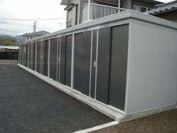 多数の引き戸が並んだ、外壁が灰色で横に細長い金属製の集合型の物置のようなリサイクル品目別の集積所が、舗装された地面の上に設置されている、本町地区ストックハウスの写真