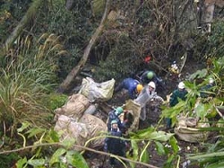 緑が生い茂る山林の斜面で、複数の作業員が軍手やヘルメットを着用して、土嚢袋やシートに包まれた大量の不法投棄物を入れたごみを、協力して撤去している作業風景を捉えた写真