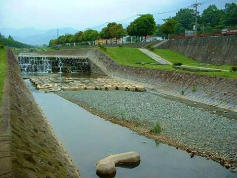 両岸がコンクリートの護岸で整備され、浅い水が流れる河川の風景で、上流には人工的な小さな段差の滝と、水面に浮きのようなものが規則的に並んでいる川の風景写真