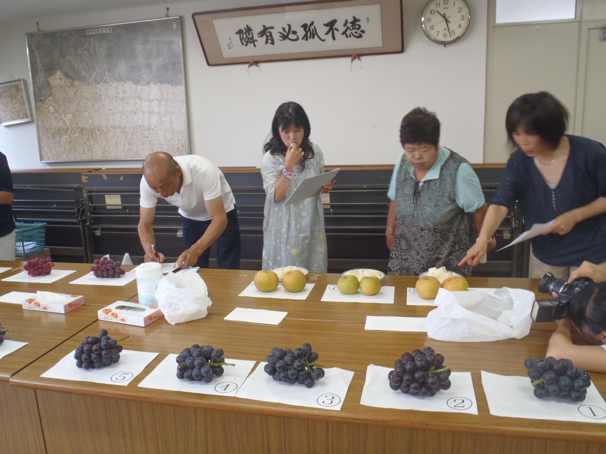 番号を振った紙の上にブドウやナシなどの果物が並べられたテーブルの前で、4人の男女が梨を指さしながら審査を行っている様子の写真