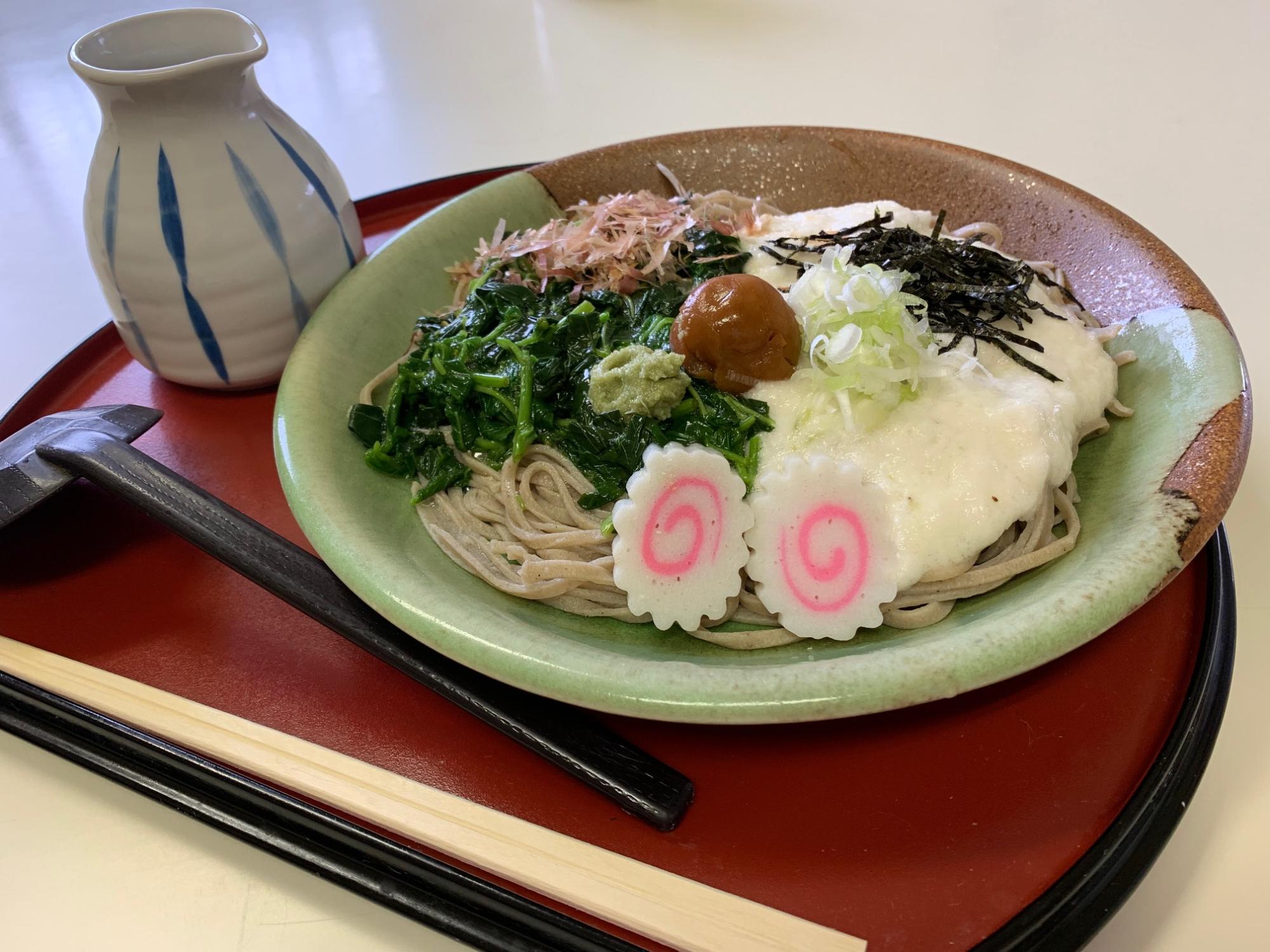 茶色と緑色の配色の皿に盛られた冷たい蕎麦の上に、とろろ、モロヘイヤ、削り節、梅干し、刻みネギ、わさび、二枚のなるとが華やかにトッピングされた夏梅とろそばの写真
