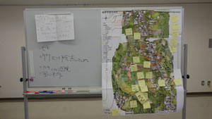 室内の壁際に置かれたホワイトボードには手書きで書かれたメモが書かれており、その横の地図には検討事項が書かれた多数の黄色い付箋が貼られている資料の写真