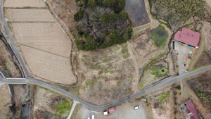 V字に交わるアスファルトの農道と水路、茶色い農地に囲まれた中央の荒れ地の、やぶ払い後の農村地域の真上からの空撮写真