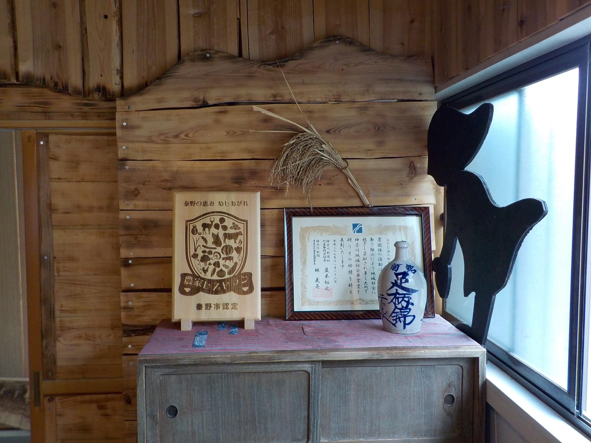木製の壁の前に置かれた古い箪笥の上に、豊野町認定の木製看板、額に入った表彰状のある農家レストラン 秦野どぶろく家の店内の写真