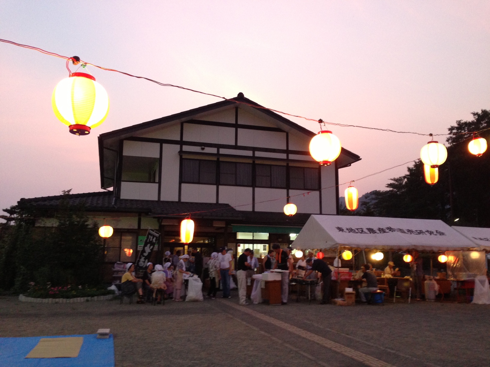 夕暮れ時のふるさと伝承館の敷地に明かりを灯した提灯が並び、建物の前の白い屋台のテントに大勢の人々が集まり賑わっている納涼まつりの様子の写真
