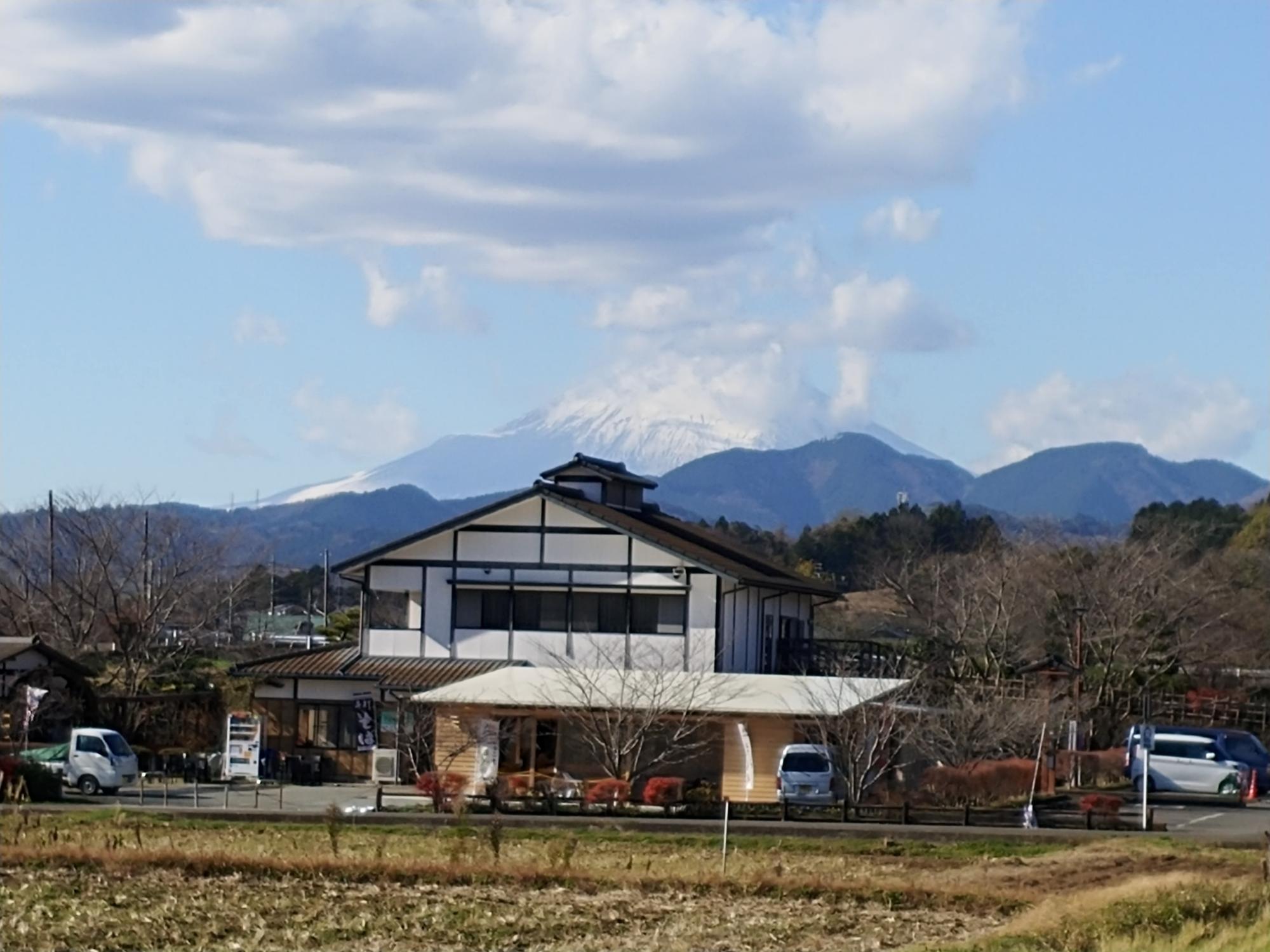 グレーの屋根に白壁の木造二階建てのふるさと伝承館の建物の背後に森が広がり、奥には雪をかぶった雄大な富士山が見える写真