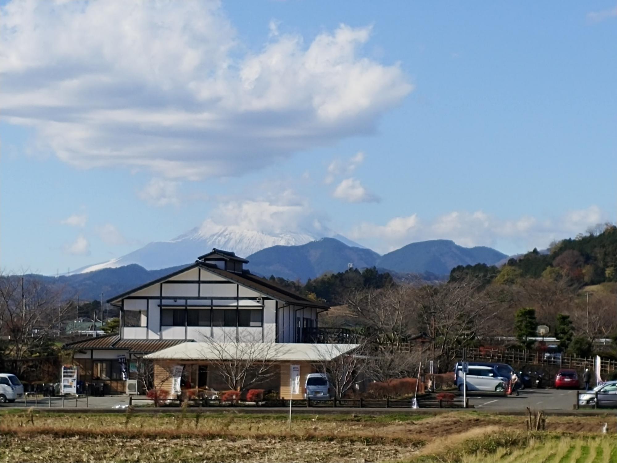 グレーの屋根に白壁の木造二階建てのふるさと伝承館の建物の背後に森が広がり、奥には雪をかぶった雄大な富士山が見える写真