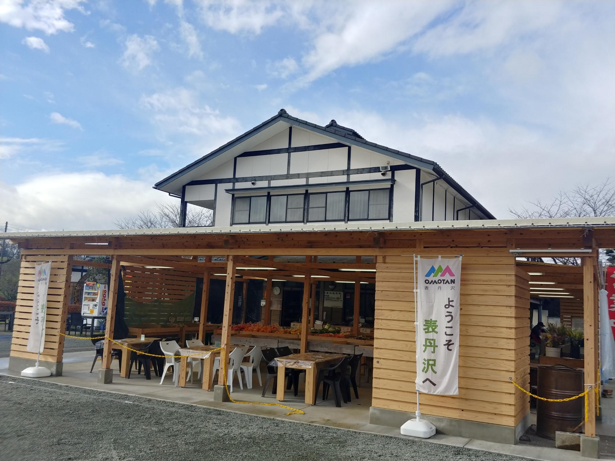 「ようこそ表丹沢へ」と書かれたのぼりが立った屋根付きのテラス内にテーブルと椅子が並び、奥では農産物が販売されている、ふるさと伝承館の前の直売所の写真