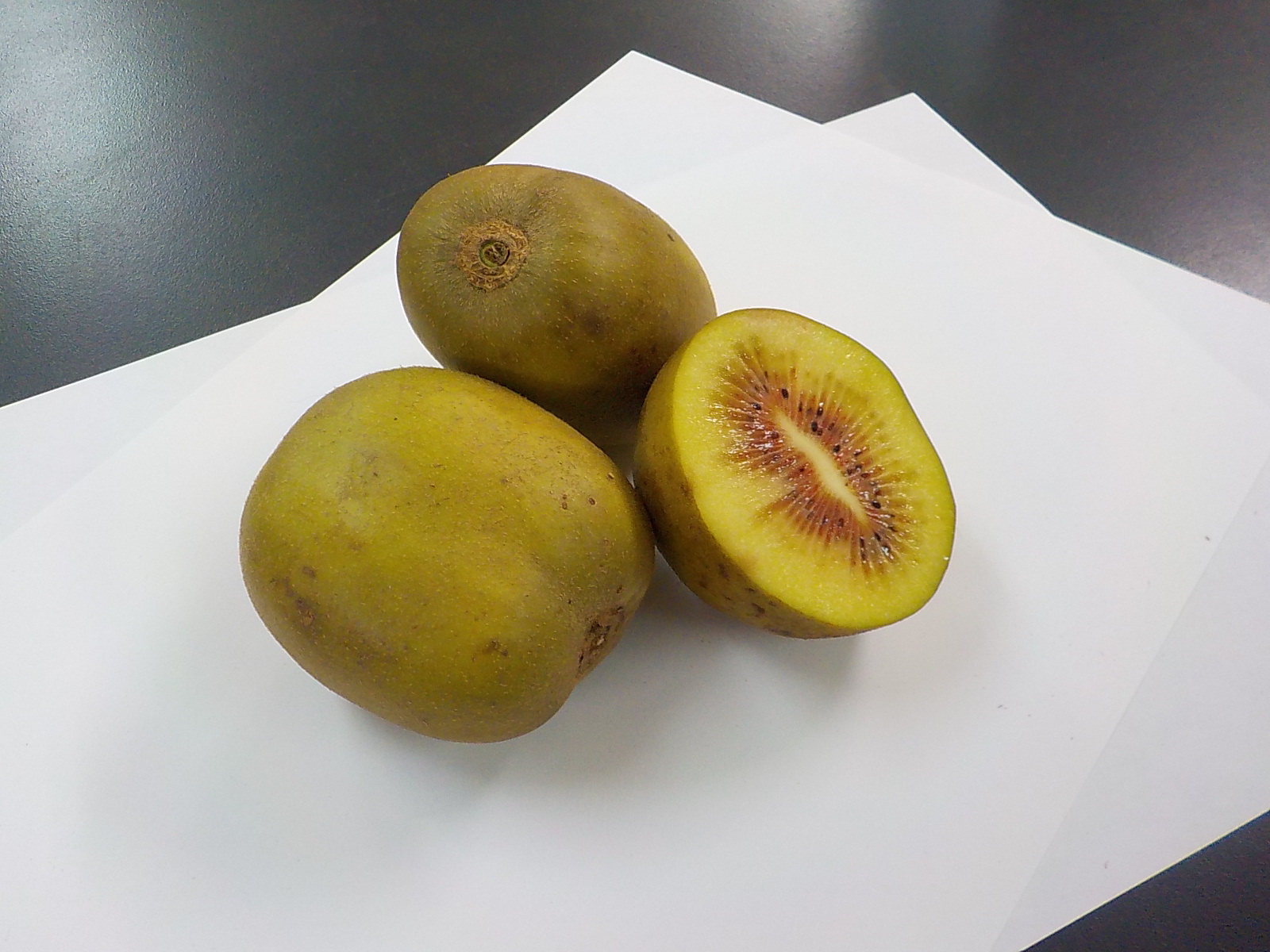 全体が茶色がかった緑色をした2個のキウイフルーツの実と、半分にカットされ黄緑色の果肉の中央に集まった種の周りが赤くなっている様子がわかるキウイフルーツが並べて置かれている写真