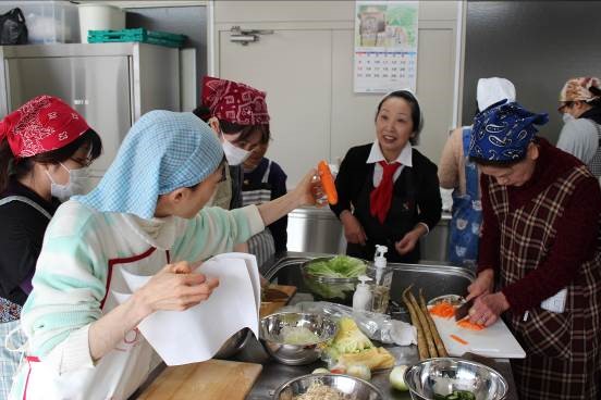 和気あいあいとした雰囲気の中で、頭にバンダナを巻いた数人の女性たちが調理実習室のような場所で、にんじんなどの野菜を切り、料理の下準備をしている様子の写真