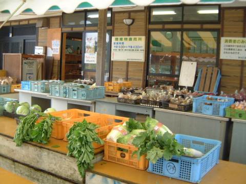 緑色のキャベツや葉物野菜、その他の農産物が入ったプラスチック製のカゴが、木造の建物前のカウンターの上に並べられている様子の写真