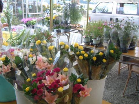 店頭のバケツやテーブルに、ピンクや黄色の花束が数多く並べられ、後方には鉢植えの植物や白いワンボックスカーが見える、屋外の生花店の様子の写真