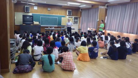 広々としたフローリングの室内で、大勢の子どもたちが床に座って前方を向き、黒板の前で立っている男性の話を聞いている様子の写真