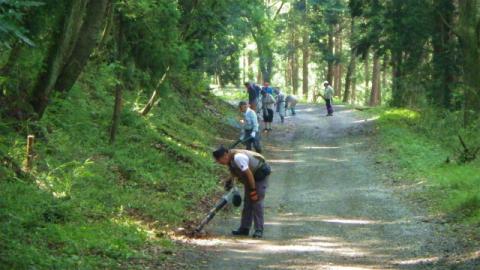 林の中の未舗装の道を、数人の人々が機材を持ち列をなして歩きながら、落ち葉の除去などの環境整備活動をしている様子の写真