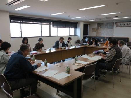 U字型に配置された会議机を囲み、資料やペットボトルのお茶が置かれ、参加者が真剣な面持ちで話し合いや資料への書き込みを行っている会議室内の写真