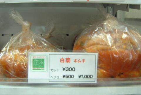 冷蔵ケースの中で、赤く漬け込まれた白菜キムチがビニール袋に詰められており、その手前には価格が書かれたポップが置かれている写真