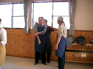 エプロンを付けた高齢の男性が仲間からエプロンをつけるのを手伝ってもらい、皆で楽しそうに笑い合っている様子の写真