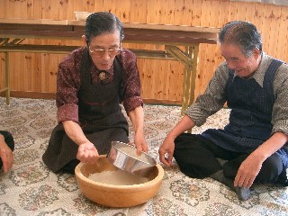 参加者が床に座り、一人の男性がが大きな木鉢の上で粉をふるい、男性がそれを見守っている、そば打ち体験様子の写真