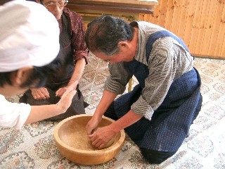そば打ち体験で、エプロンをつけた高齢の男性が木鉢の中の生地を一生懸命こねており、周りの仲間がそれを見守っている様子の写真