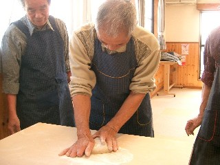 そば打ち体験で、エプロンをつけた高齢の男性がのし台の上で力強く生地をこね、もう一人の男性がその様子を真剣に見守っている写真