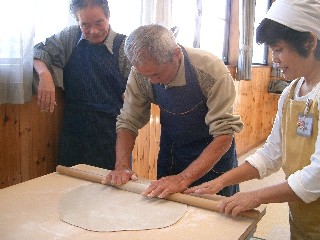 そば打ち体験で、女性が補助しながら、高齢の男性が麺棒で生地を薄く伸ばしており、その様子を別の男性が笑顔で見守っている写真