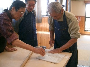 そば打ち体験で、他の参加者に教わりながら高齢の男性が麺切り包丁で生地を細く切り、別の男性が後ろから見守っている様子の写真