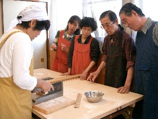 そば打ち教室で、講師が麺切り包丁で生地を切る手本を見せており、周りでは4人の受講生が真剣な表情でその手元を覗き込んでいる写真