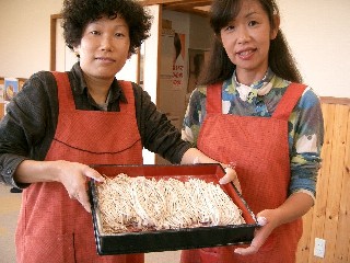 そば打ちを終えたエプロン姿の女性2人が、できあがったばかりの瑞々しい生そばを盆に乗せ、誇らしげにカメラに見せている様子の写真
